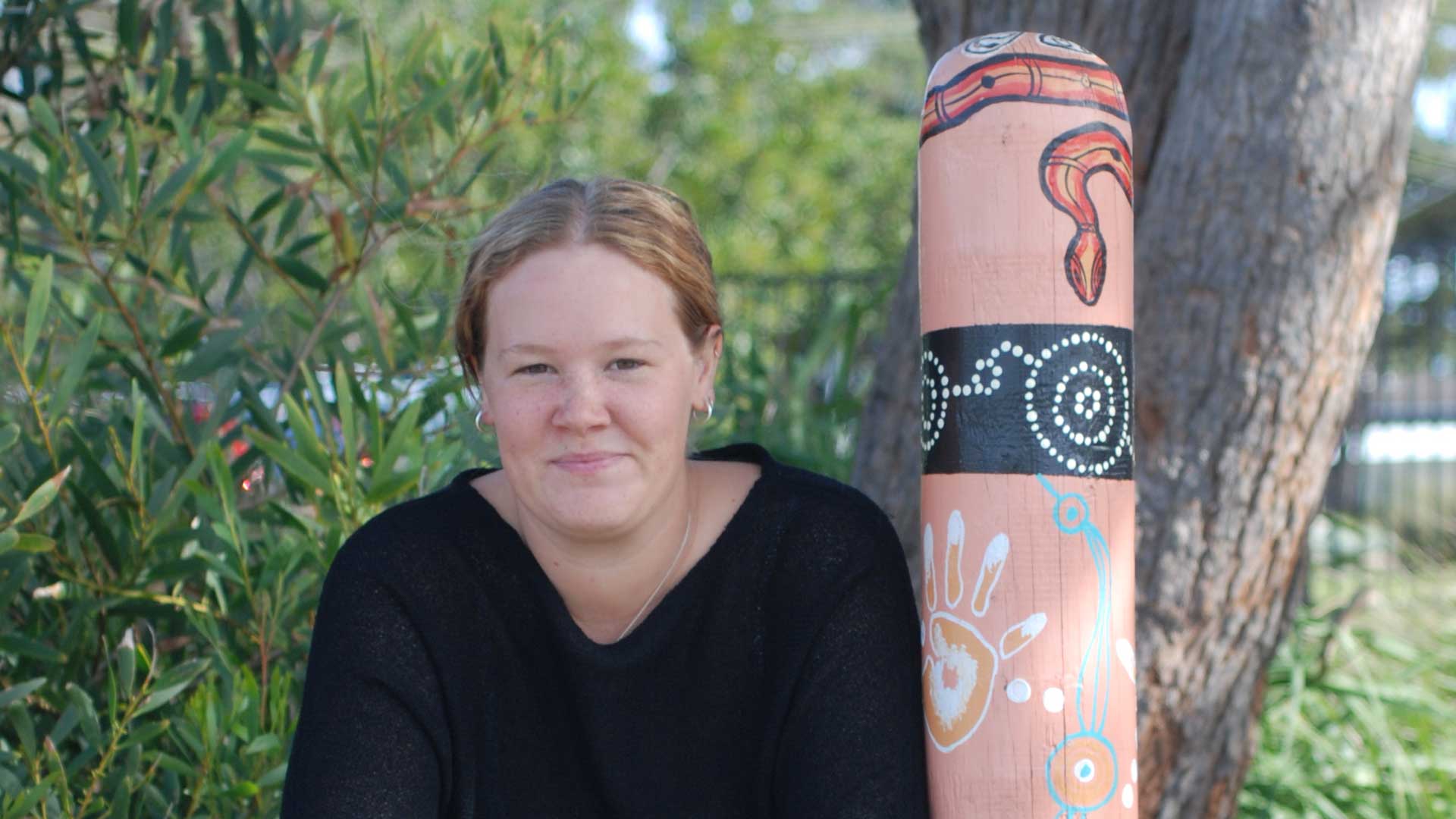 Photo of Takesha Frank smilink next to Aboriginal artwork