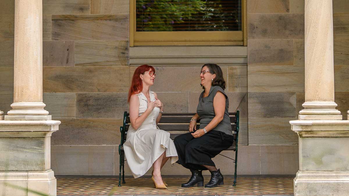 two women talking on a bench outside a historical stone building