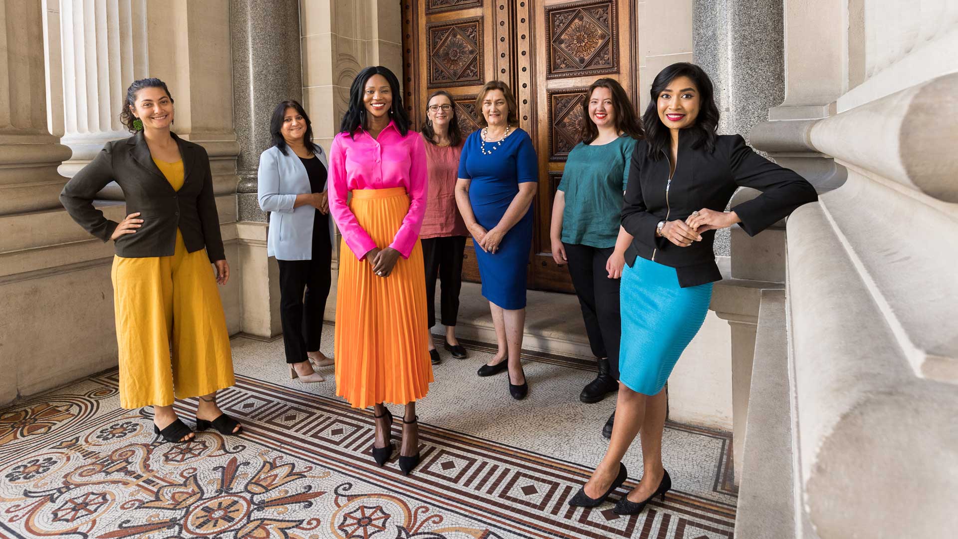 7 smiling women wearing smart, brightly coloured clothes, posing against sandstone pillars