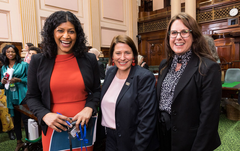 Three smiling women posing for a photo in the Victorian Parliament chamber: Samantha Ratnam MLC, Juliana Addison MP and Cathrine Burnett-Wake MLC.