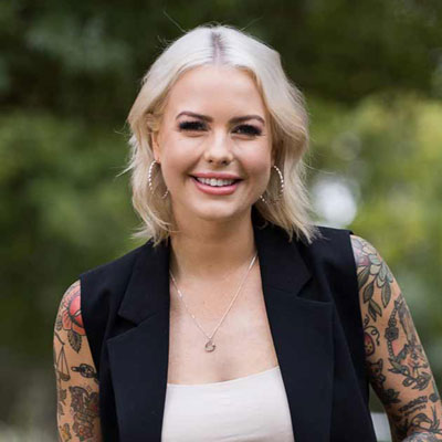 Photo of Georgie Purcell smiling, bare arms covered in tattoos, greenery in the background