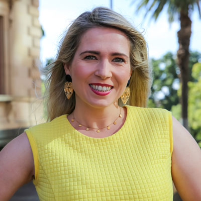 Photo of Jenna Davey-Burns smiling wearing a bright yellow summer dress, historical building and palm tree in the background