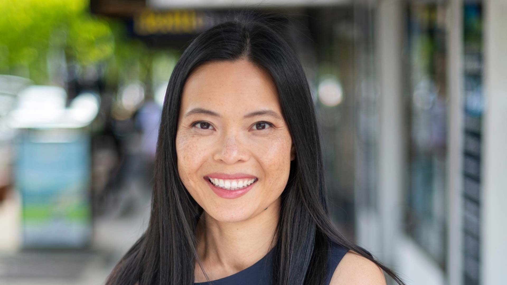 Photo of Sally Sitou MP smiling, suburban shopping strip in the background