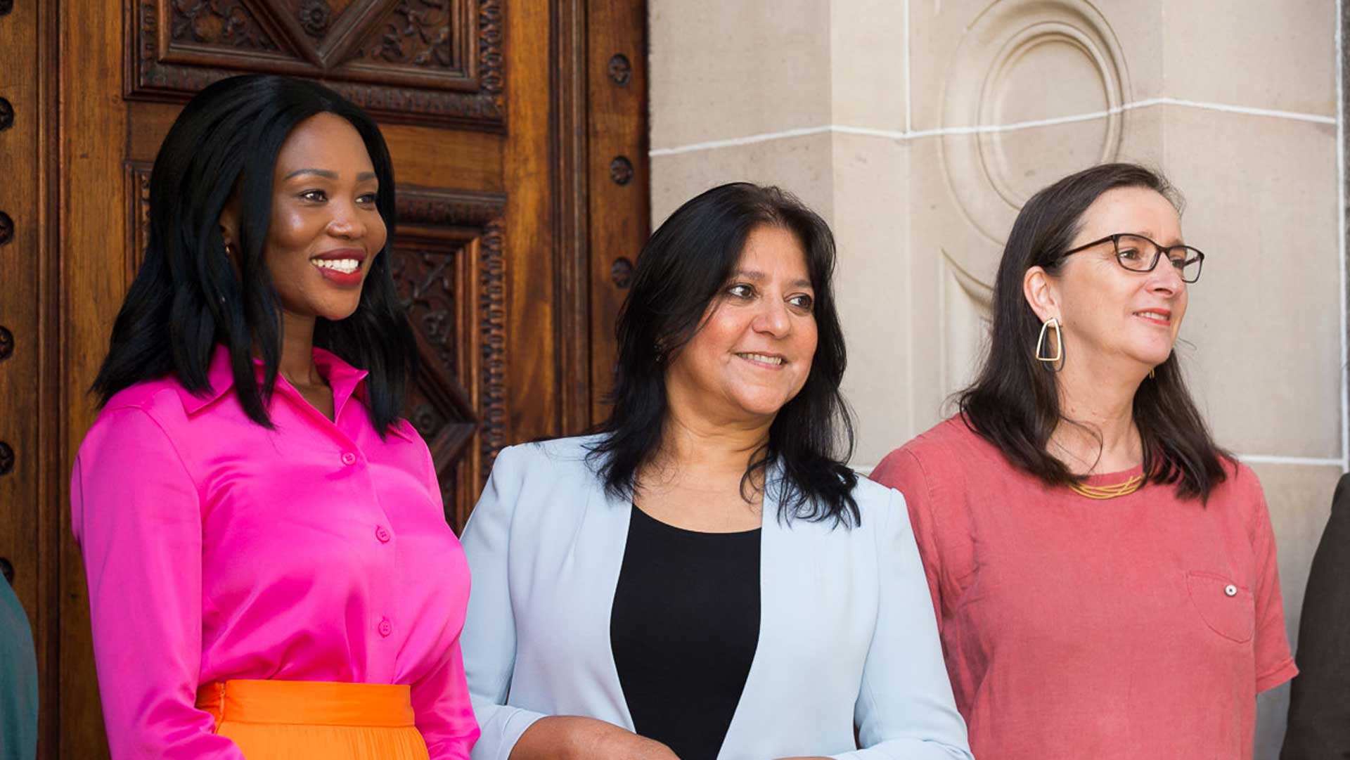 Candid, smiling photo of three brightly dressed diverse women standing outside a parliamentary building