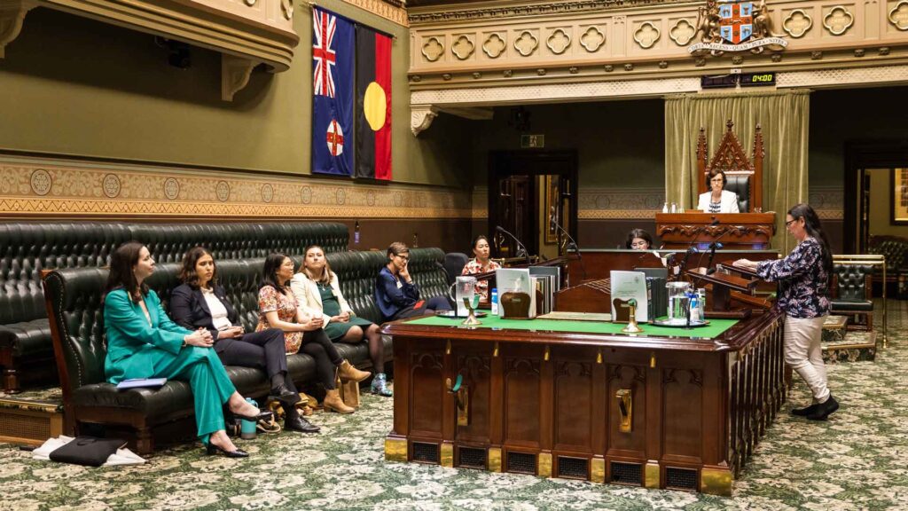 Photo of a small group of women inside the NSW parliament chamber, Australian and Aboriginal flags in the background