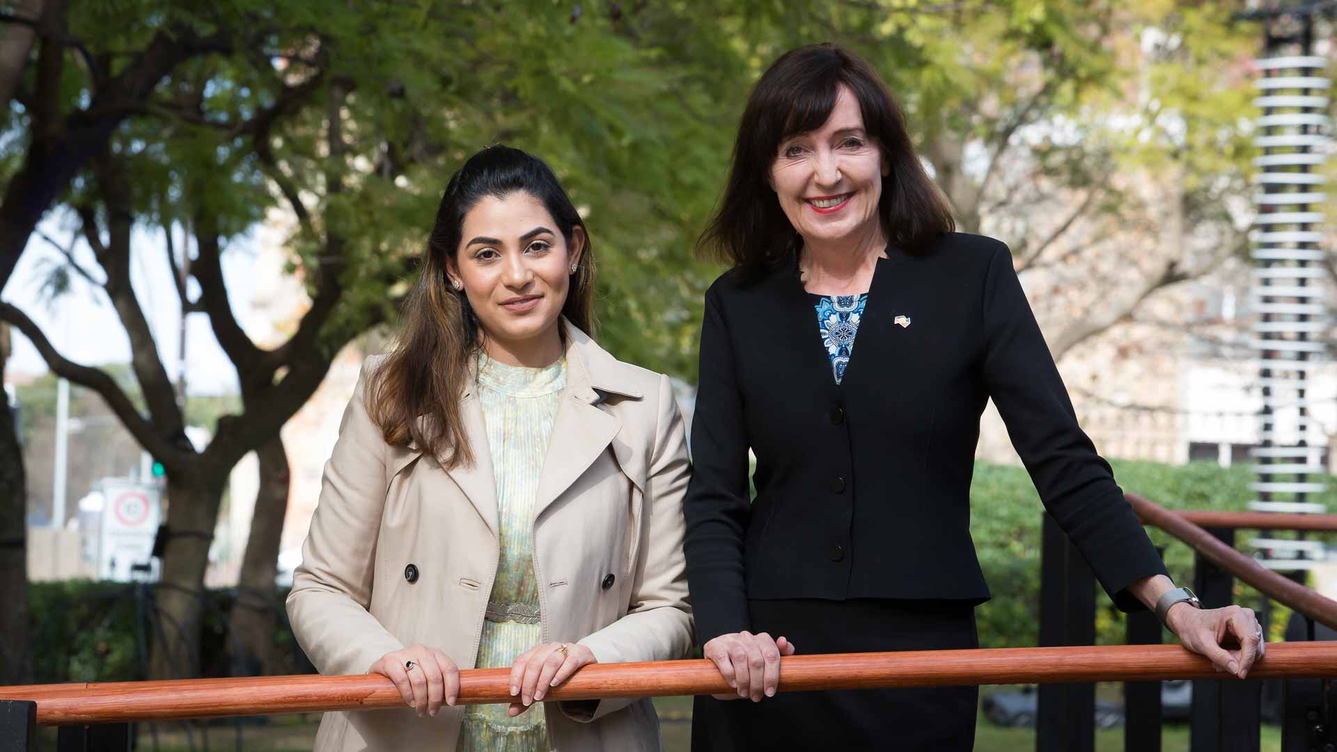 A woman in a navy suit and a younger woman in beige jacket pose smiling in front of trees and greenery.