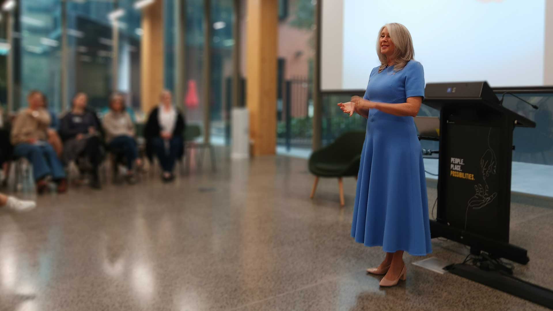 Tracey Spicer wearing a blue dress standing in front of a large screen, delivering to an audience of women