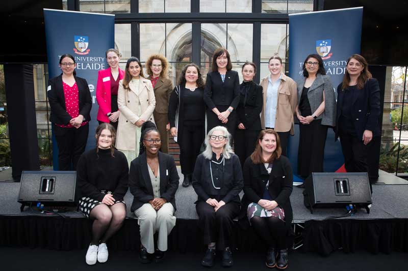 A group of 14 smiling women of different ages and cultural backgrounds pose on a stage, four of them sitting on the edge of the stage. There are two dark blue banners with the University of Adelaide logo behind the group.