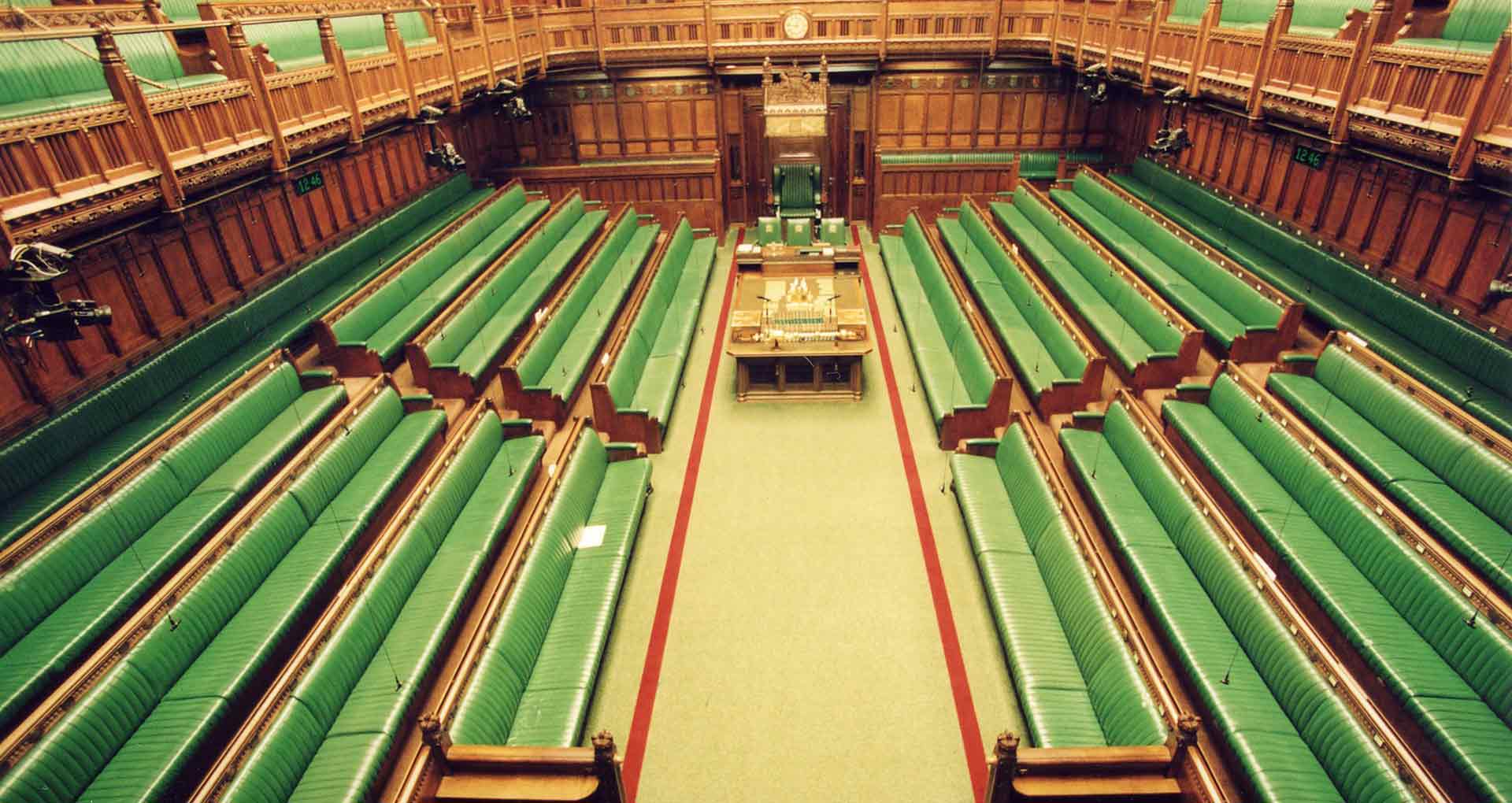 Photo of the inside of the UK House of Commons showing 8 rows of empty green leather bench seats and dark wood panelling on the walls
