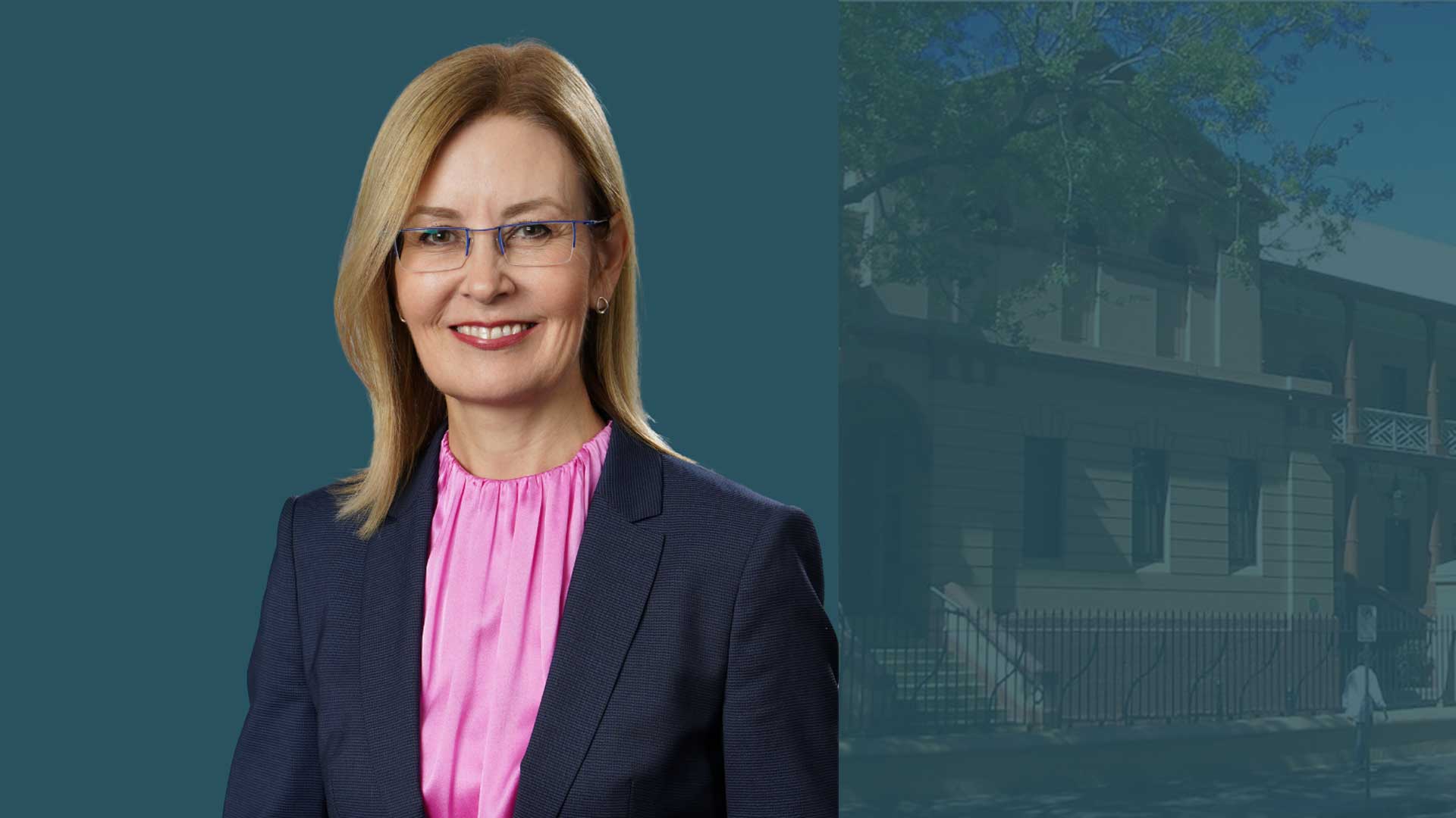 Photo of Gabrielle upton in a navy blazer and pink blouse. She has long fair hair and glasses. Behind her is a blue background and image eof the NSW parliament building.