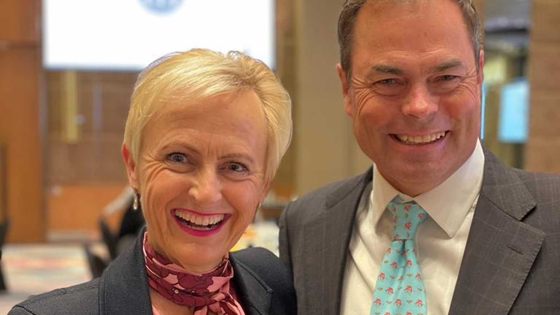 Photo of Katie and Malcolm Allen smiling. Katie has short blond hair and is wearing a grey blazer and burgundy and pink floral scarf. Malcolm is wearing a grey blazer and turquoise tie with a white shirt.