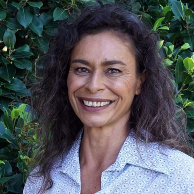 Photo of councillor Moo D'Ath smiling against a background of green foliage. She has curly dark hair and is wearing a lightly patterned shirt.