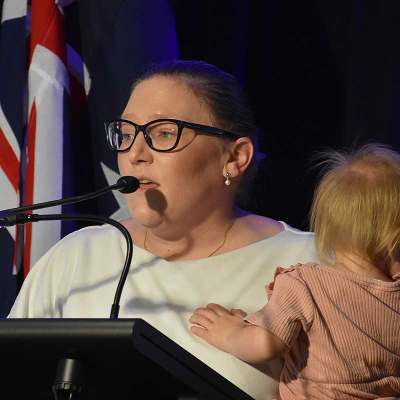 Woman wearing glasses and a cream coloured blouse speaking into a podium microphone while holding a fair-haired toddler. The Australian flag can be seen in the background.