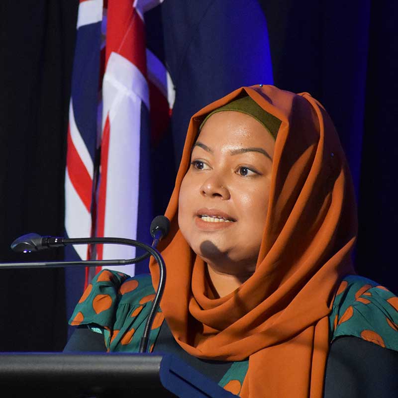 Woman wearing a rust coloured satin headscarf speaking into a podium microphone. The Australian flag can be seen in the background.