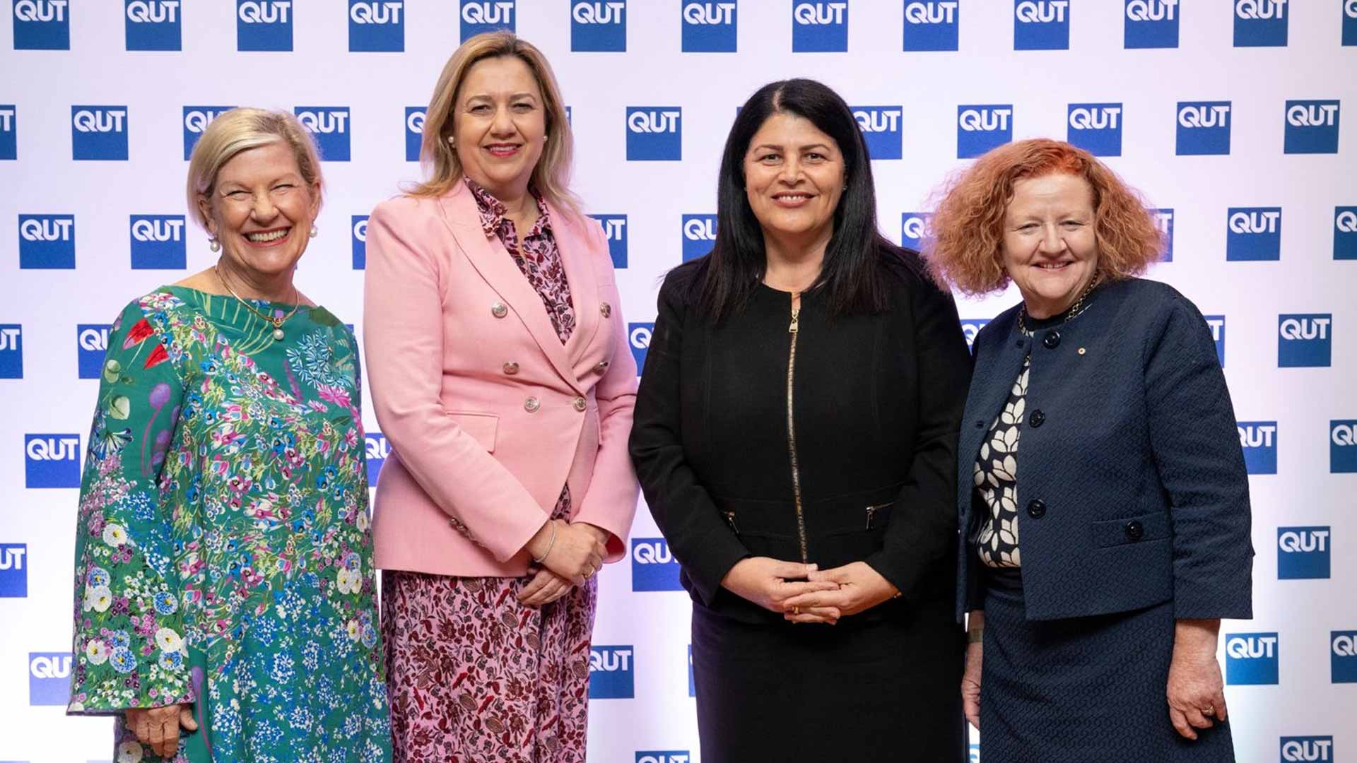 Four smartly dressed women pose in front of a media wall with the blue QUT repeated across it.