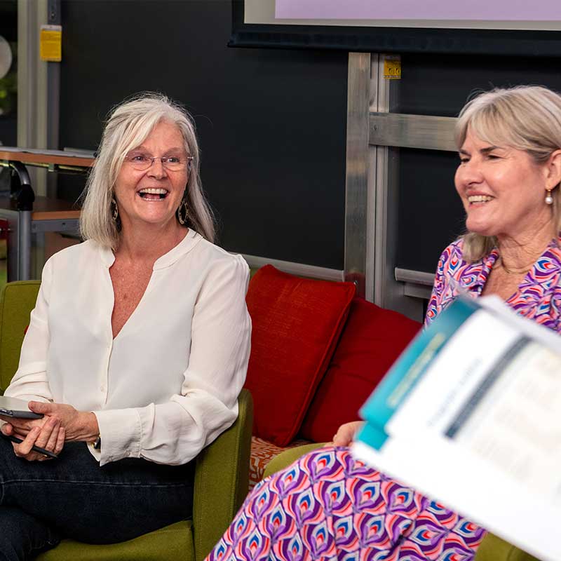 Two women seated in padded red chairs, talking and smiling. One has long grey hair and a white blouse. The other has shorter grey hair and a patterned red dress.