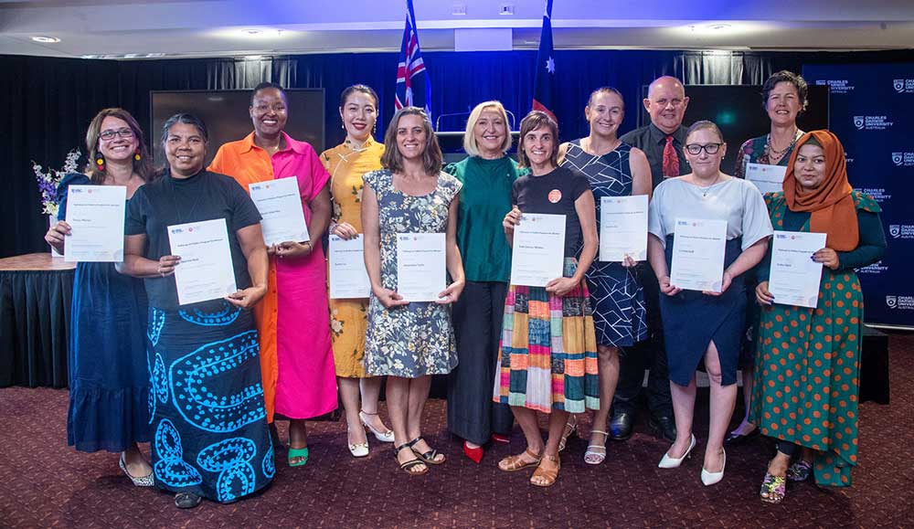 A group of colourfully dressed and culturally diverse women pose smiling, holding white graduation certificates. The Australian flag can be seen in the background.