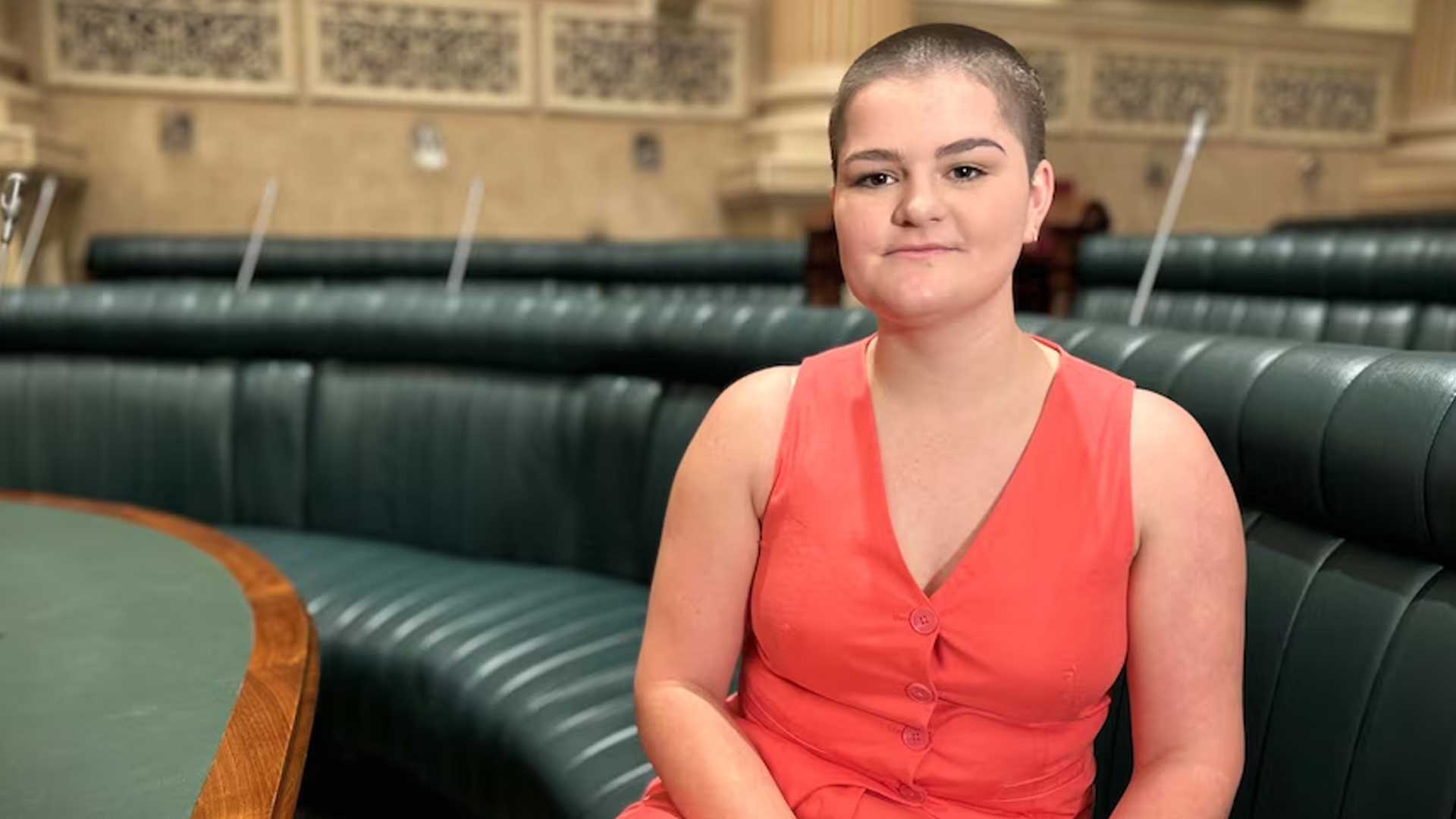 Photo of Sallee Shepherd, a young woman with close cropped hair wearing a red short sleeved jumpsuit. she is sitting in a parliamentary chamber on curved green bench seating.