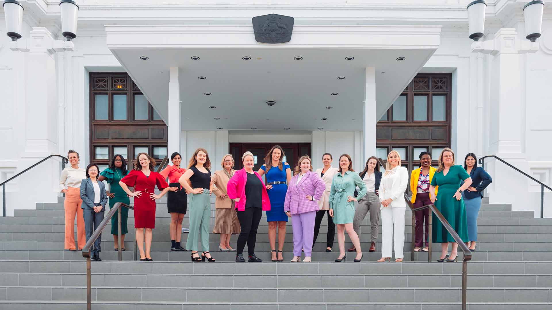 Large group of diverse women, wearing colourful professional clothing, standing on the steps of Canberra's Old Parliament House.