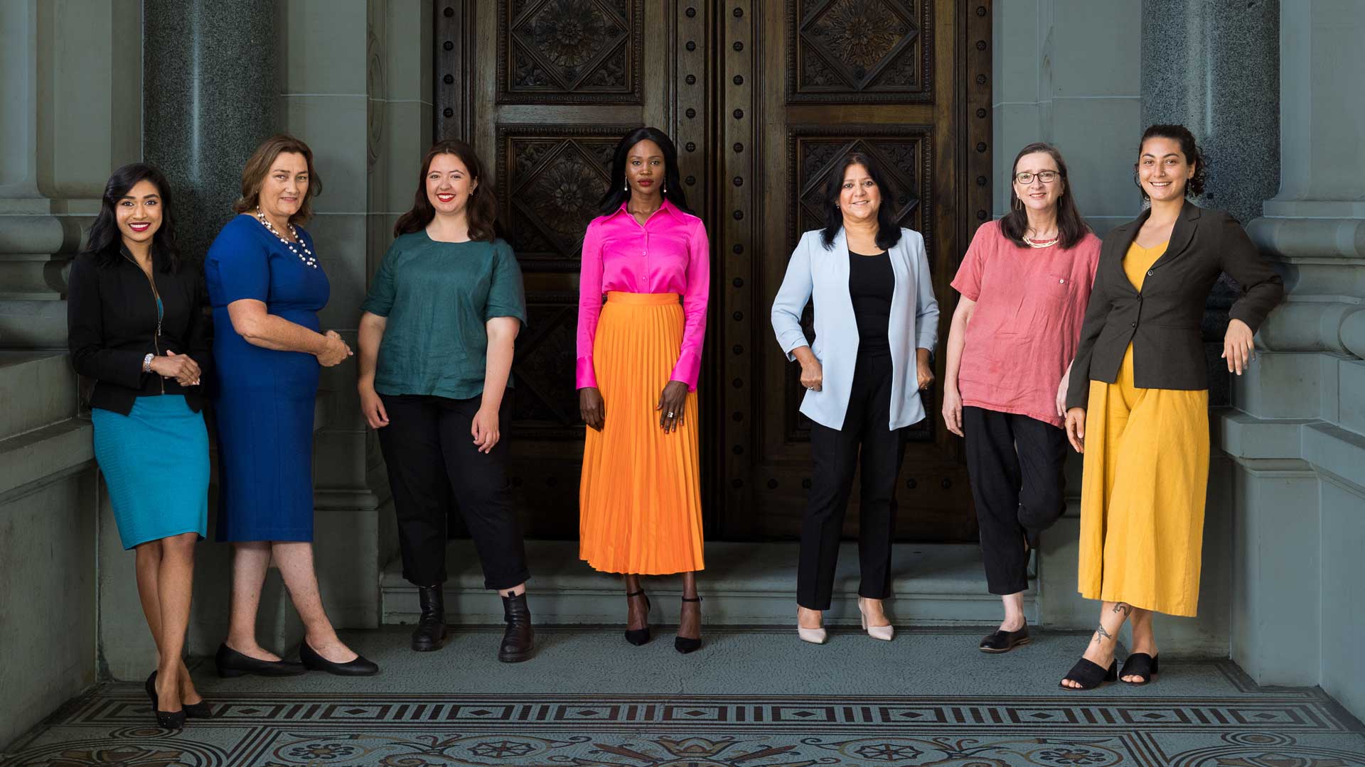 Group of 7 diverse women wearing bright block coloured clothing. the background is an exterior of a historic parliamentary building, washed out in dark blue, making the women stand out against the background.