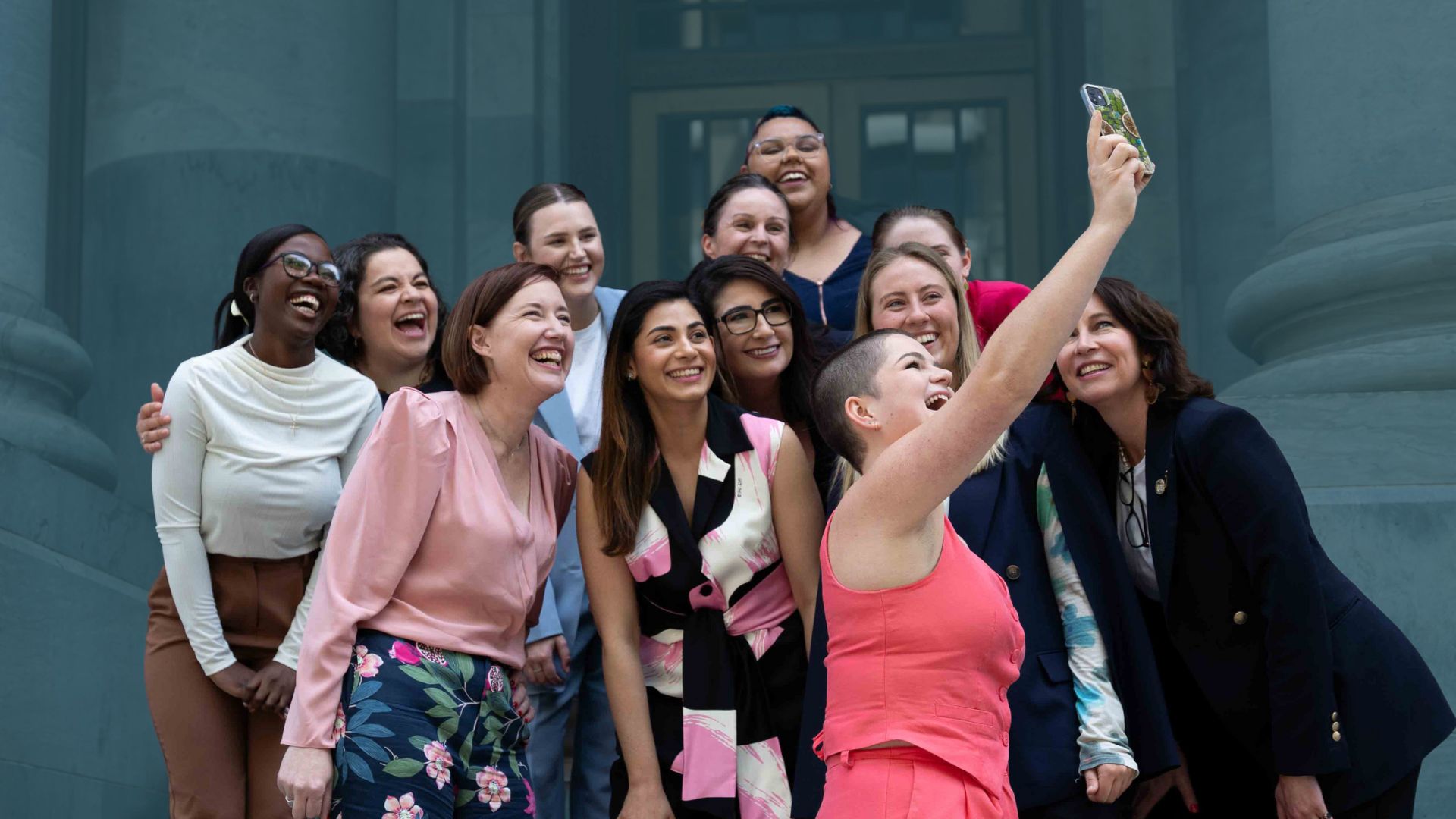 A group of 12 diverse women standing outside a historical parliament building. They are smiling as a young woman in their group takes a selfie of the group.
