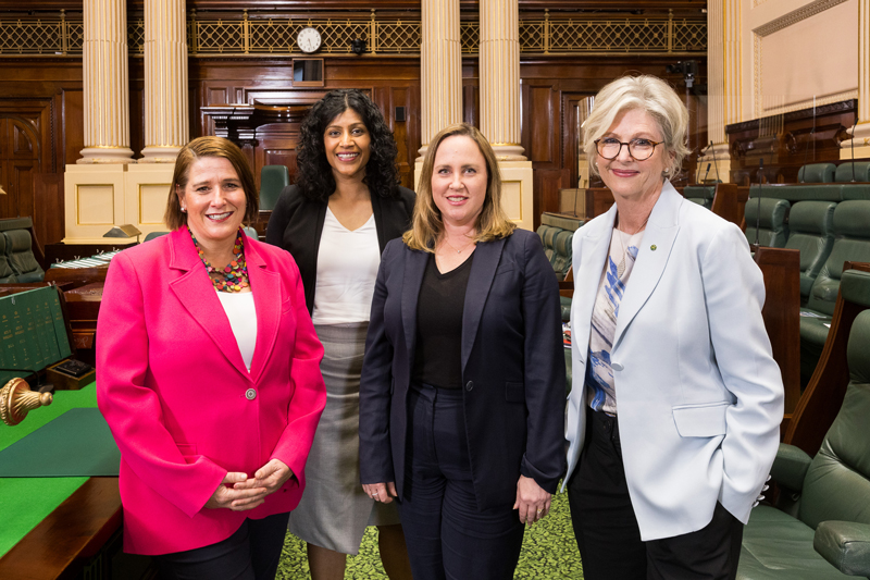 Four women stand posing in parliamentary chamber with green carpet and wooden panelling.