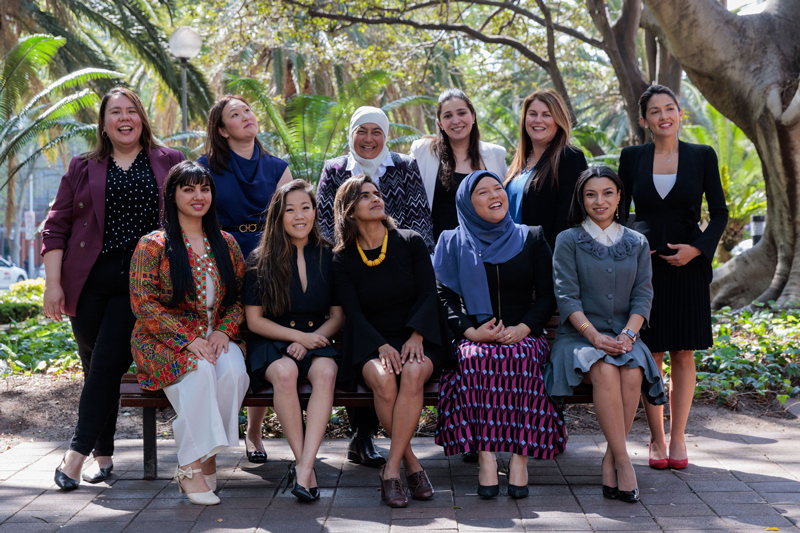 A group of 11 diverse women smile and pose for a photo outside, trees in the background.