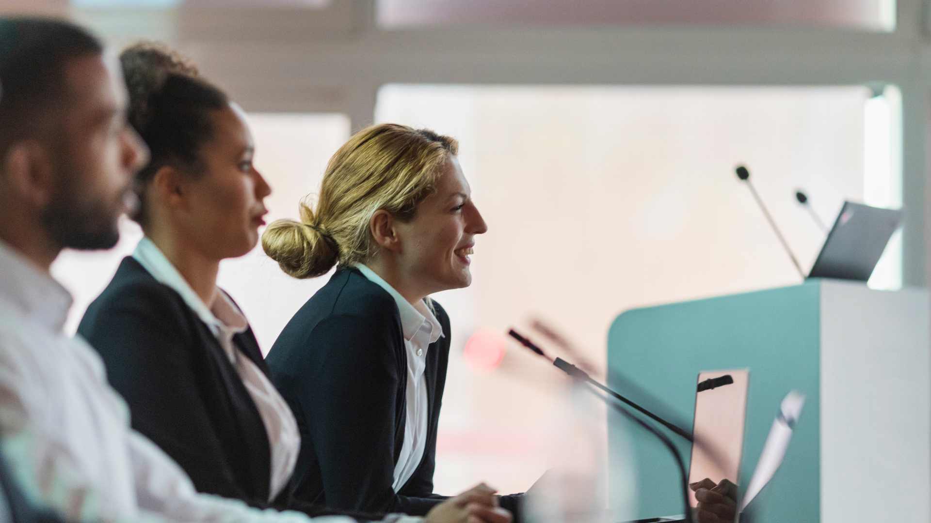 Photo of two women in navy blazers leaning forward to speak into microphones in a small conference room setting