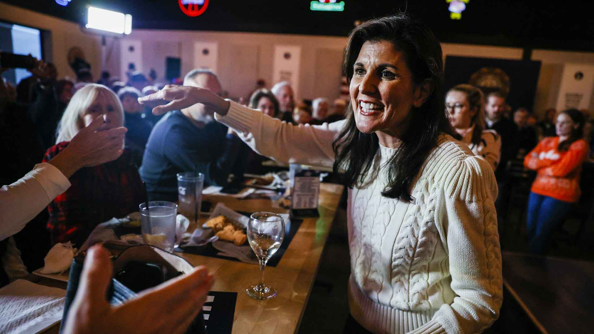 Photo of US presidential candidate Nikki Haley, a middle aged woman with long brown hair, smiling and wearing a cream coloured jumper, in a busy bar