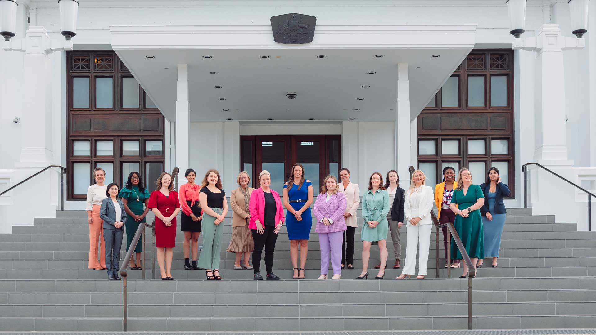 Photo of 17 diverse women wearing colourful business dress, standing on the steps of a parliamentary building