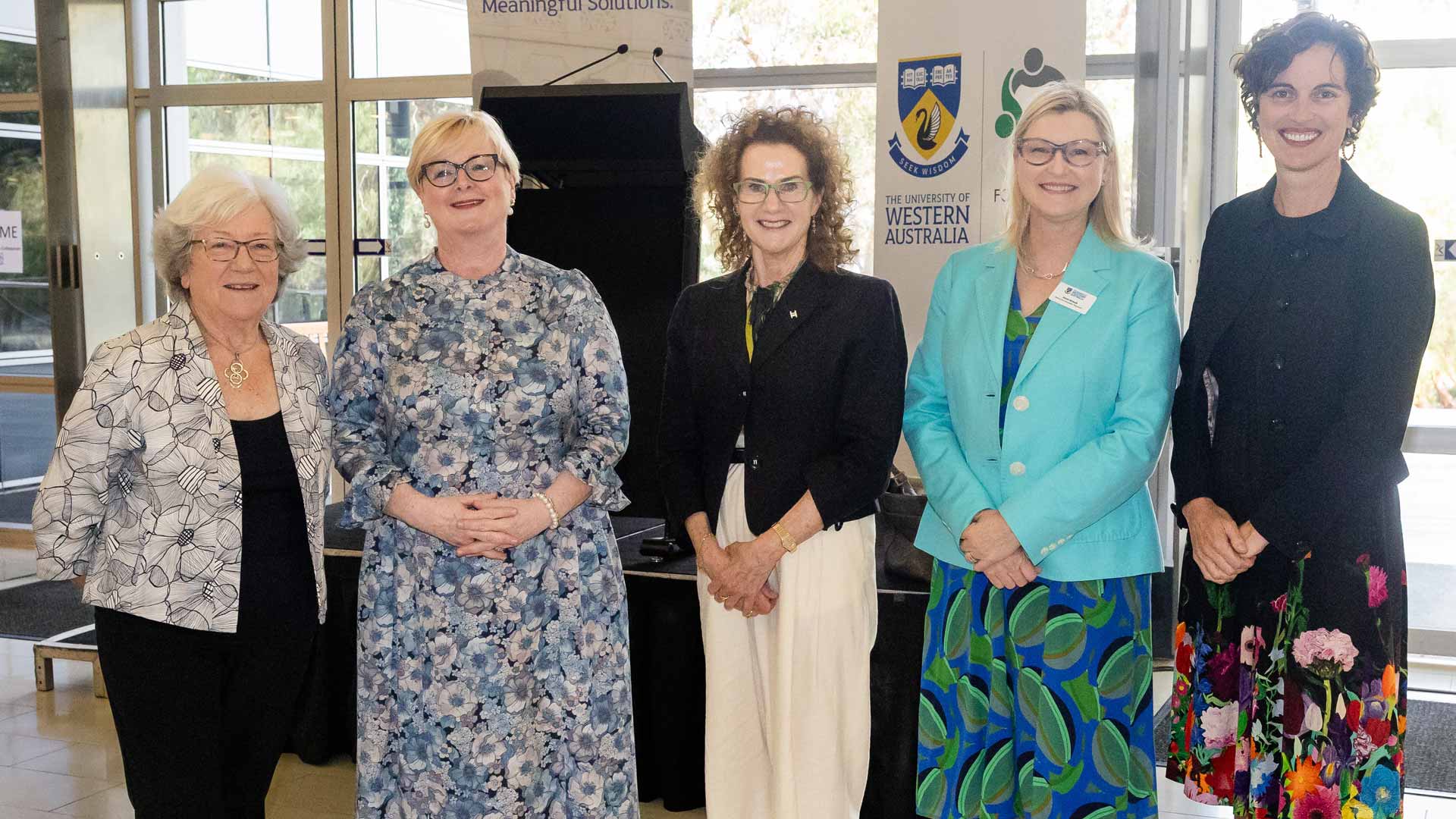 Photo of five women in colourful business dress posing for a photo in front of a podium and banner advertising The University of Western Australia