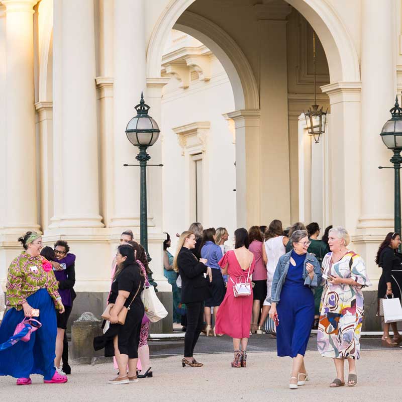 Large group of women talk in groups outside the pillars of Government House