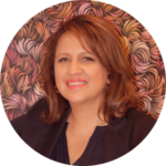 Photo of Kat Henaway smiling. She is wearing a black blazer and shirt and is standing against autumnal coloured patterned background