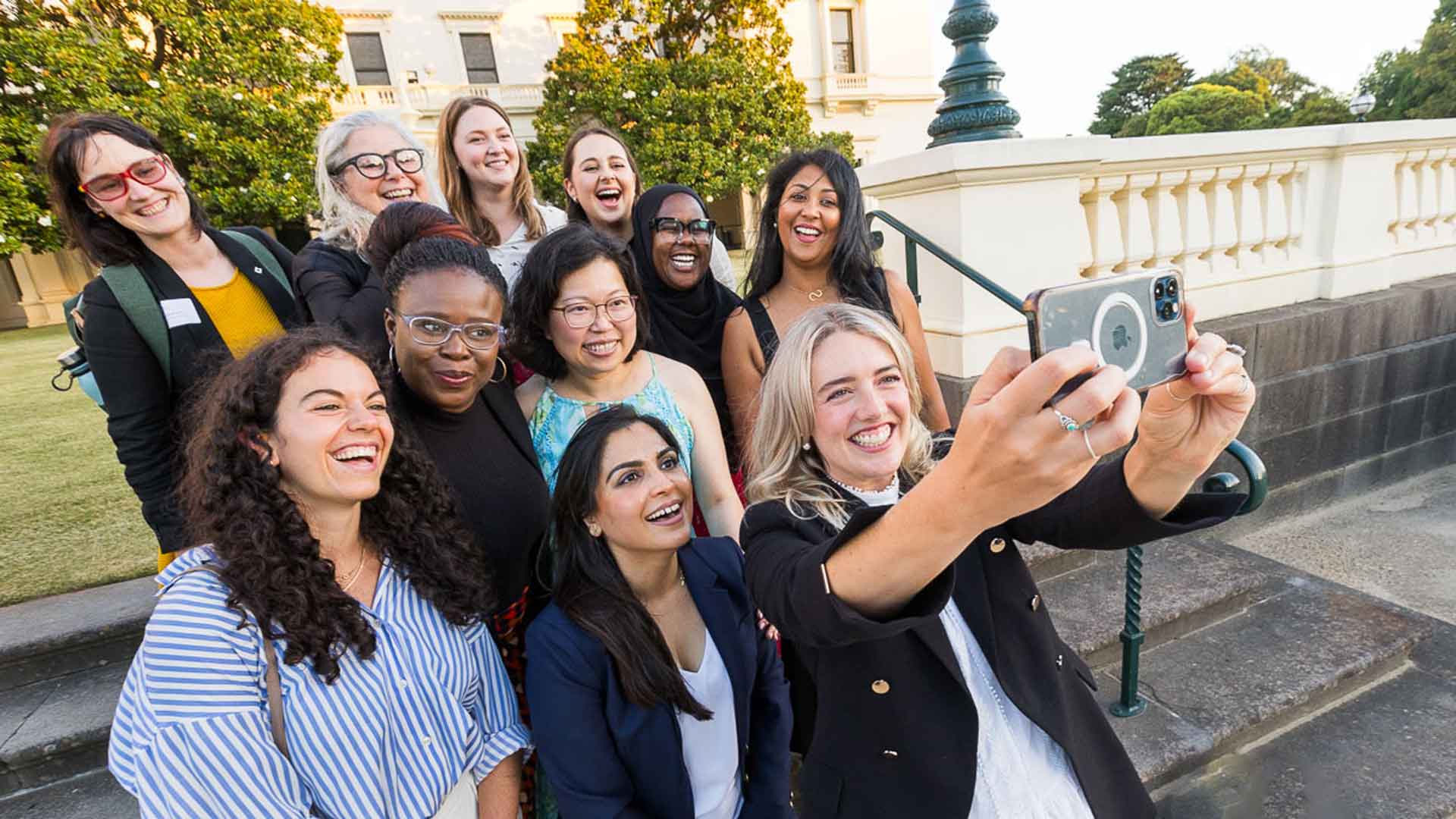 Group of 11 culturally diverse women smiling outside Victoria's government house building and lawn. One of them has her phone held up to take a selfie of the group.