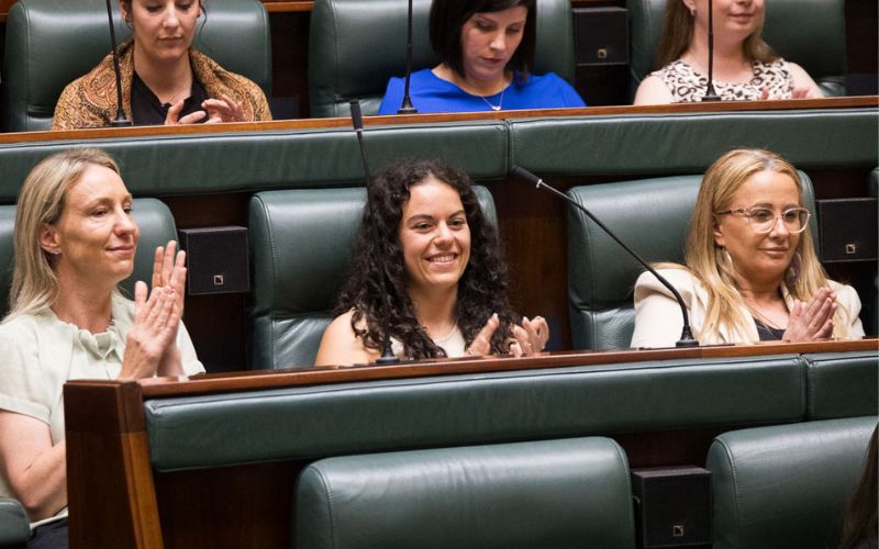 Photo of Angelica di Camillo, a young white woman with curly dark hair. She is clapping and smiling, seated in parliament