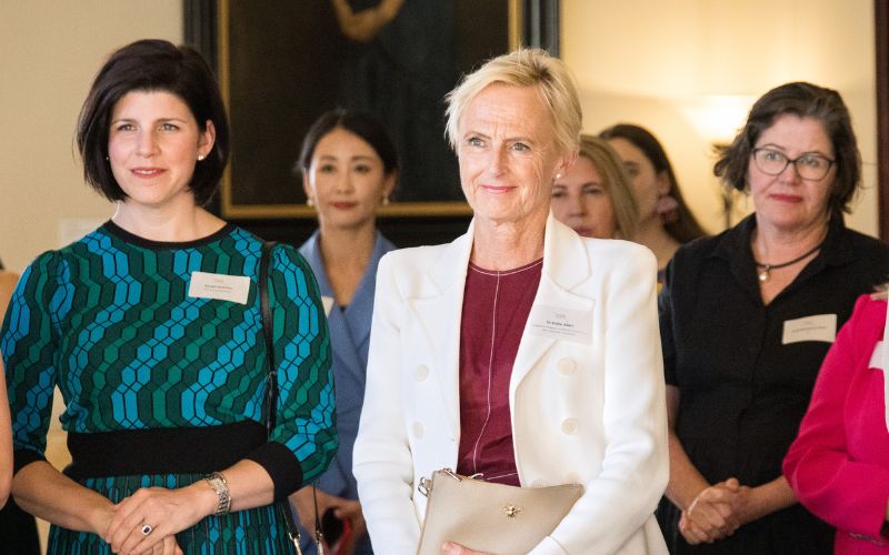 Photo of Katie Allen, a white woman with short blonde hair wearing a white suit, standing in a formal room with other women,