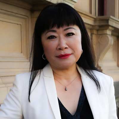 Photo of Karen Kim, a woman of Asian appearance with long black hair, wearing a white blazer, standing in front of a Victorian stone building