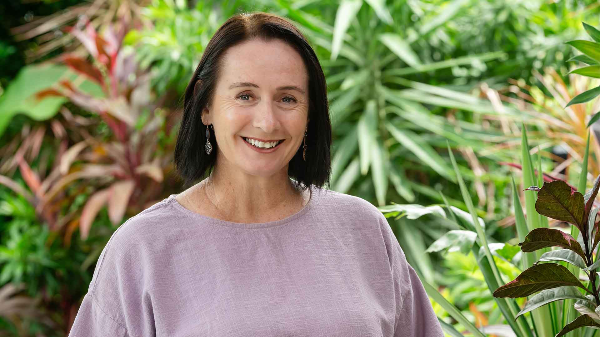 Photo of Laurie Zio, a white woman with straight dark hair, wearing a pale purple blouse. There is tropical greenery in the background.