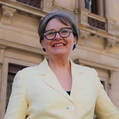 Photo of Susan Benedyka, a white woman with short blonde hair, wearing glasses and a pale yellow blazer, standing in front of a Victorian stone building