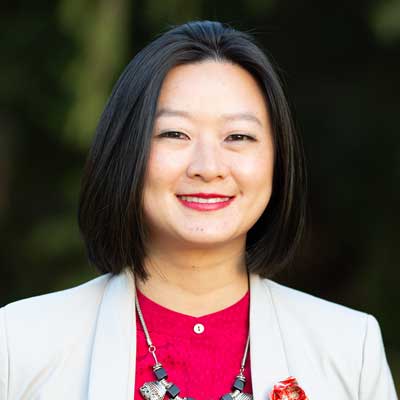 Photo of Wesa Chau, a woman of Asian appearance with dark bobbed hair, wearing a white blazer over a red blouse
