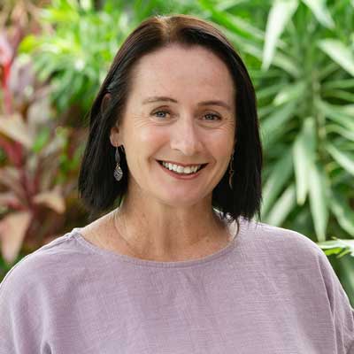 Photo of Laurie Zio, a white woman with straight dark hair, wearing a pale purple blouse. There is tropical greenery in the background.