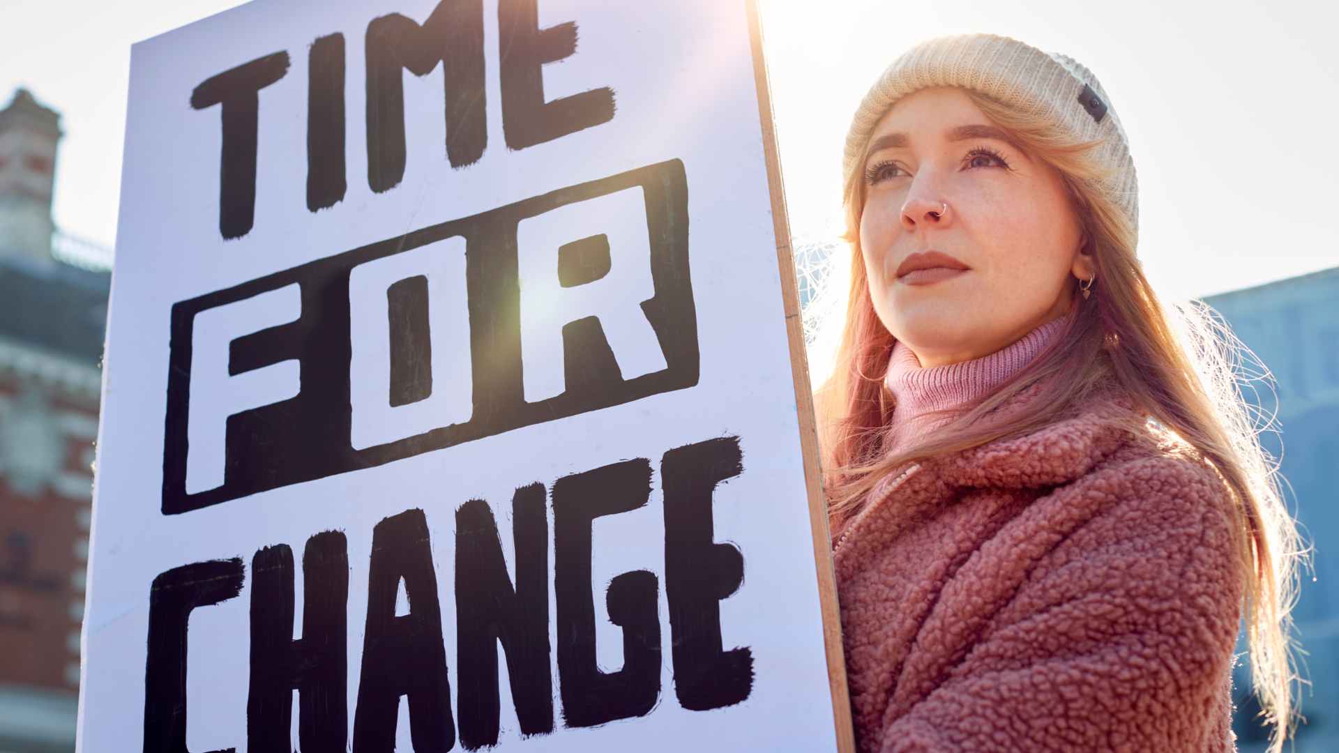 Woman in beanie and warm coat standing outside holding a sign reading TIME FOR CHANGE