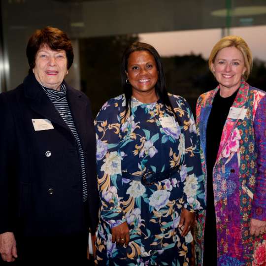 Photo of three diverse smiling women. Twi are wearing bright floral outfits, one is in a black pantsuit.