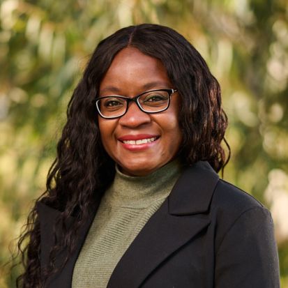 Photo of Soëlily Consen-Lynch, a black woman wearing glasses and a black blazer, smiling, trees in the background