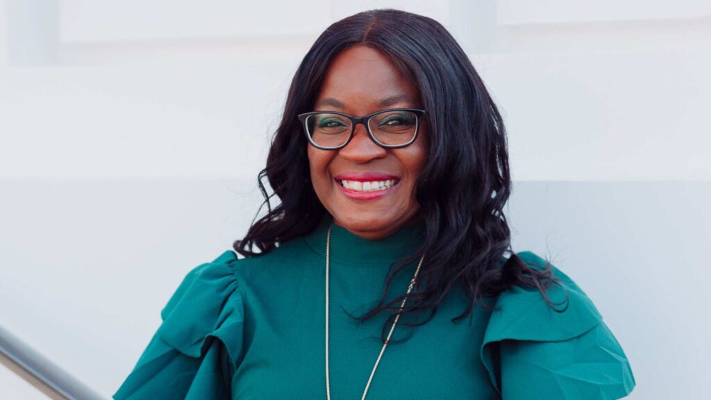 Photo of Soelily Consen Lynch smiling outside a stone building. She is a black woman wearing glasses and a teal green dress with ruffled shoulders.