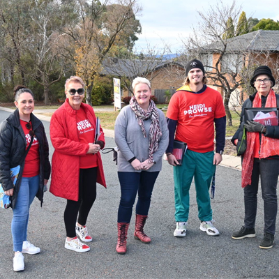 5 people standing and smiling in the road. They are dressed for cold weather and 3 of them are wearing red "Heidi Prowse" t-shirts