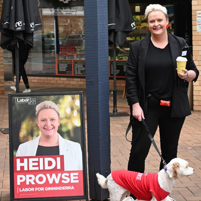 Photo of Heidi Prowse smiling. She is wearing all black, standing next to a placard of herself, and holding a small dog on a lead.