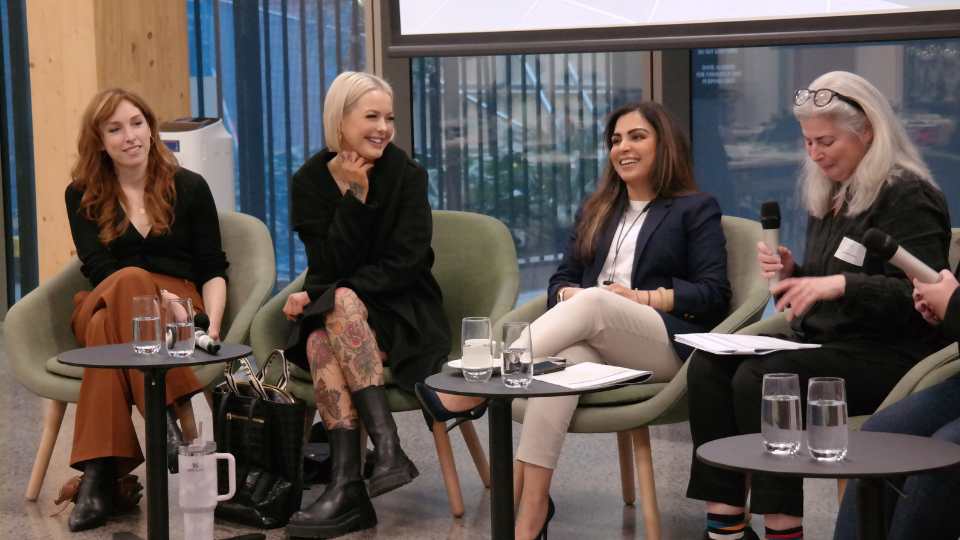 Four women sitting panel-style in low chairs, holding microphones, laughing in conversation