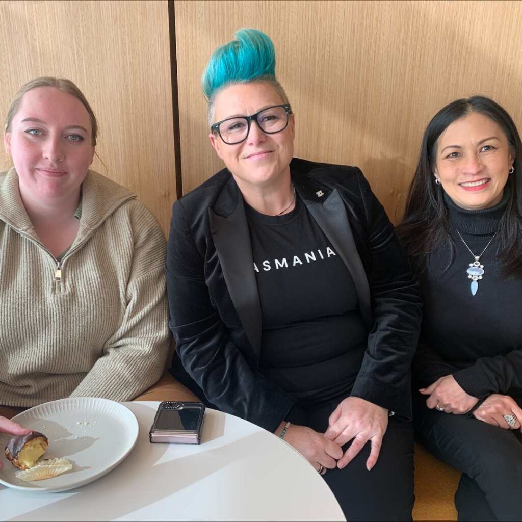 Photo of Katy Cooper, a white woman with bright blue hair and glasses, sitting at a table with two other smiling women