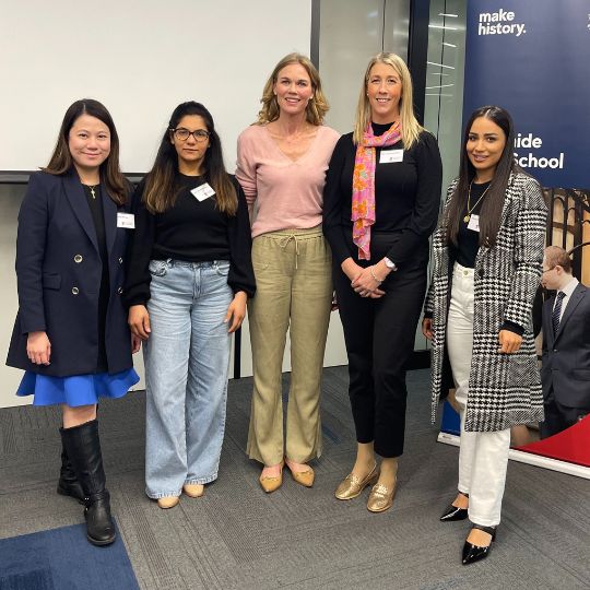 Five diverse women pose for a photo in front of a University of Adelaide banner